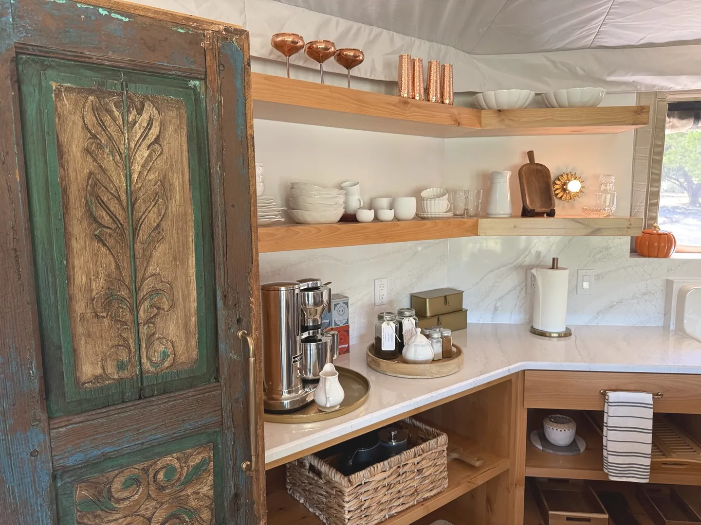 A kitchen counter area featuring open wooden floating shelves, white dishware, a coffee maker, and an ornate distressed wooden door.
