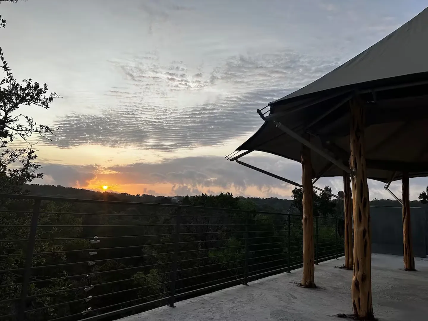 Elevated outdoor patio overlooking a dense tree canopy during sunset, with a tented canopy supported by rustic wood posts.