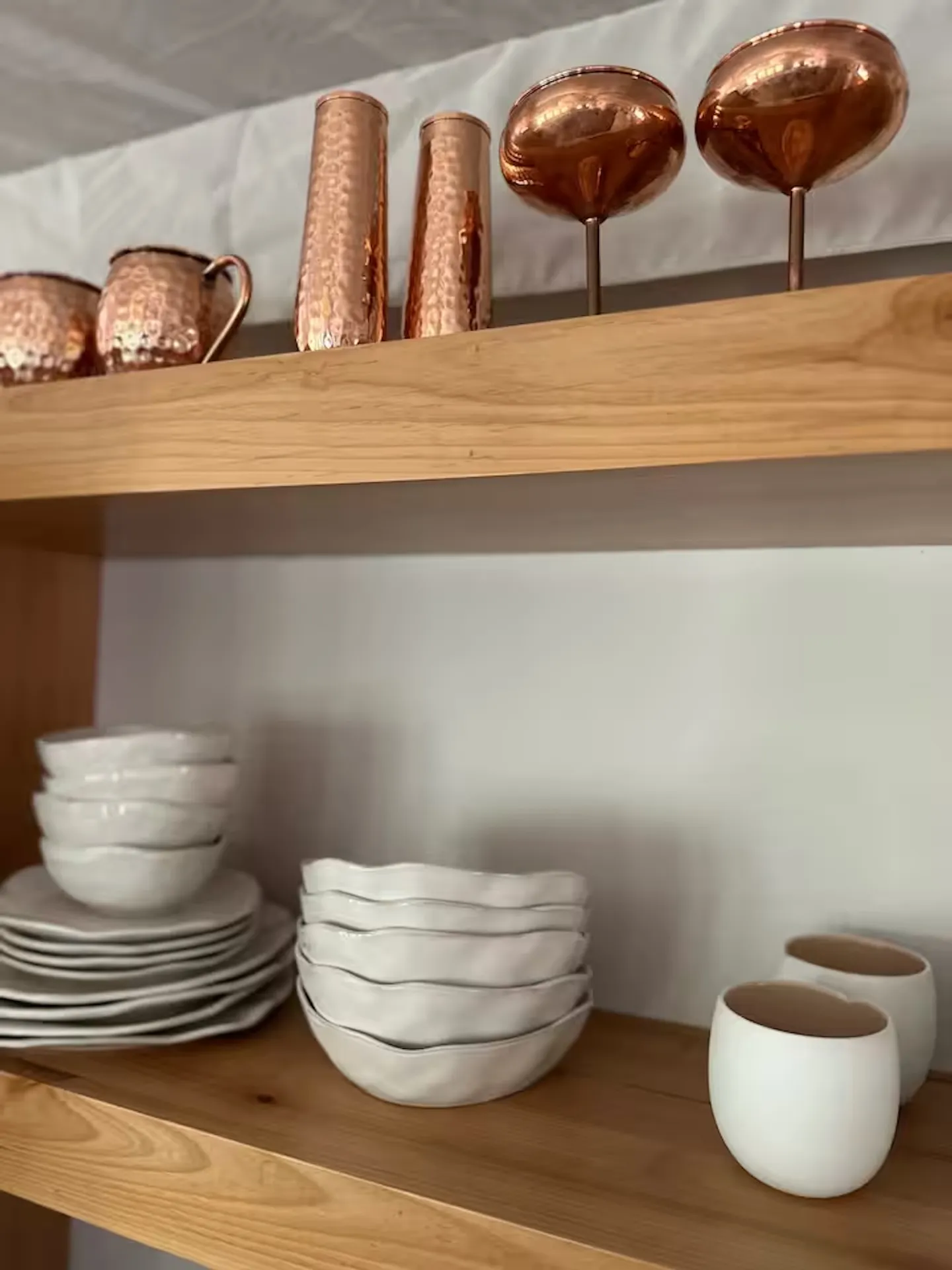 Open wooden shelves holding white ceramic dishware, mugs, and hammered copper drinkware.