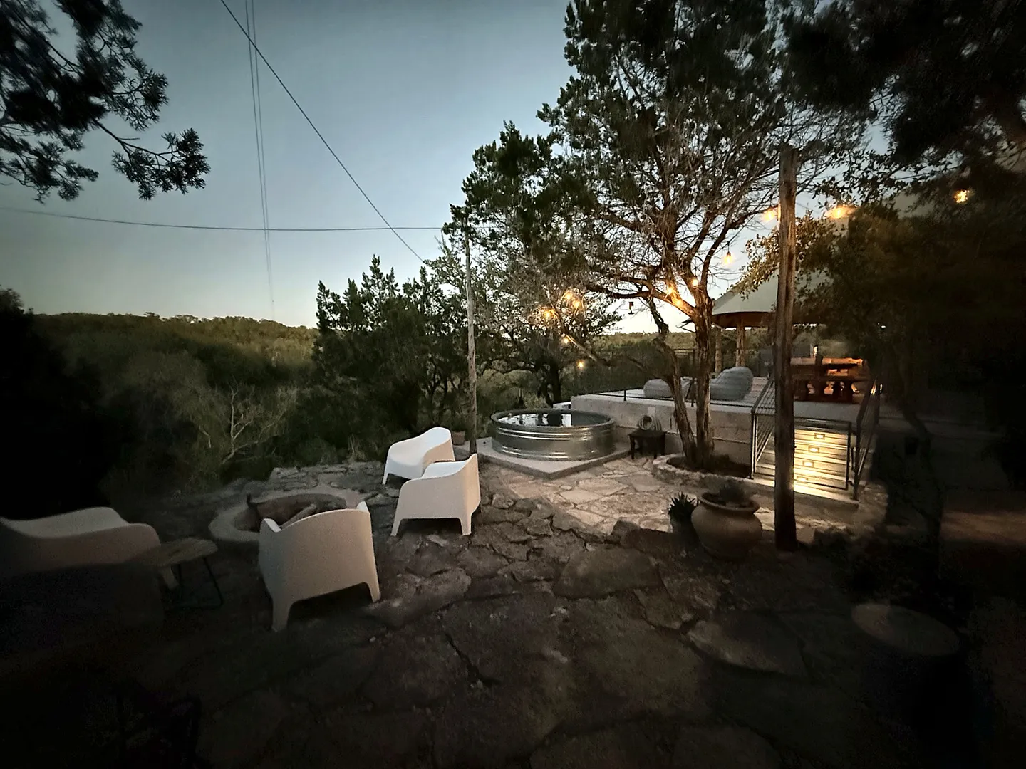 Dusk view of an outdoor stone patio with modern white chairs around a fire pit and a stock tank pool overlooking dense trees.