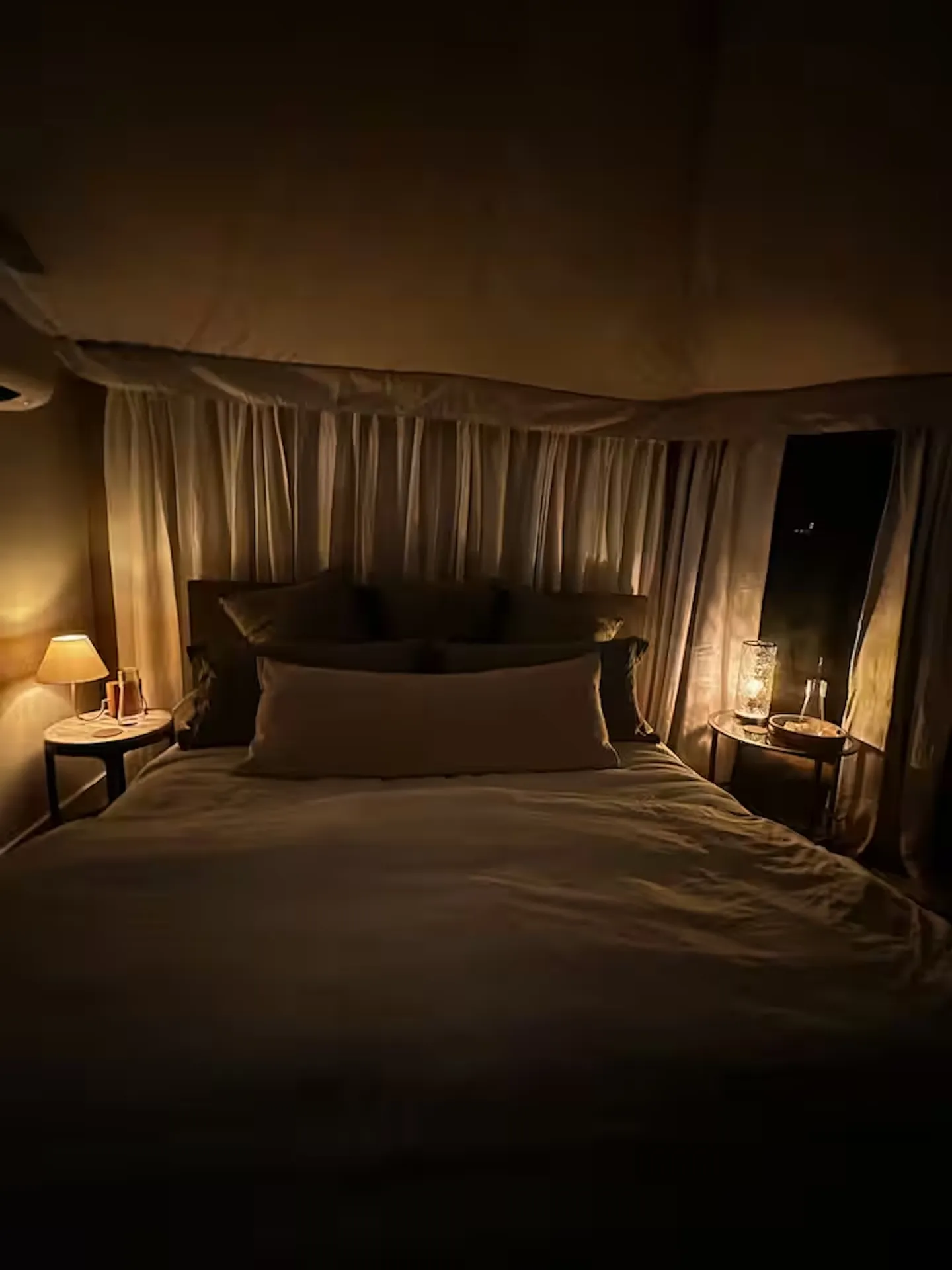 A made bed illuminated by bedside lamps at night inside a tented bedroom structure with draped curtains.
