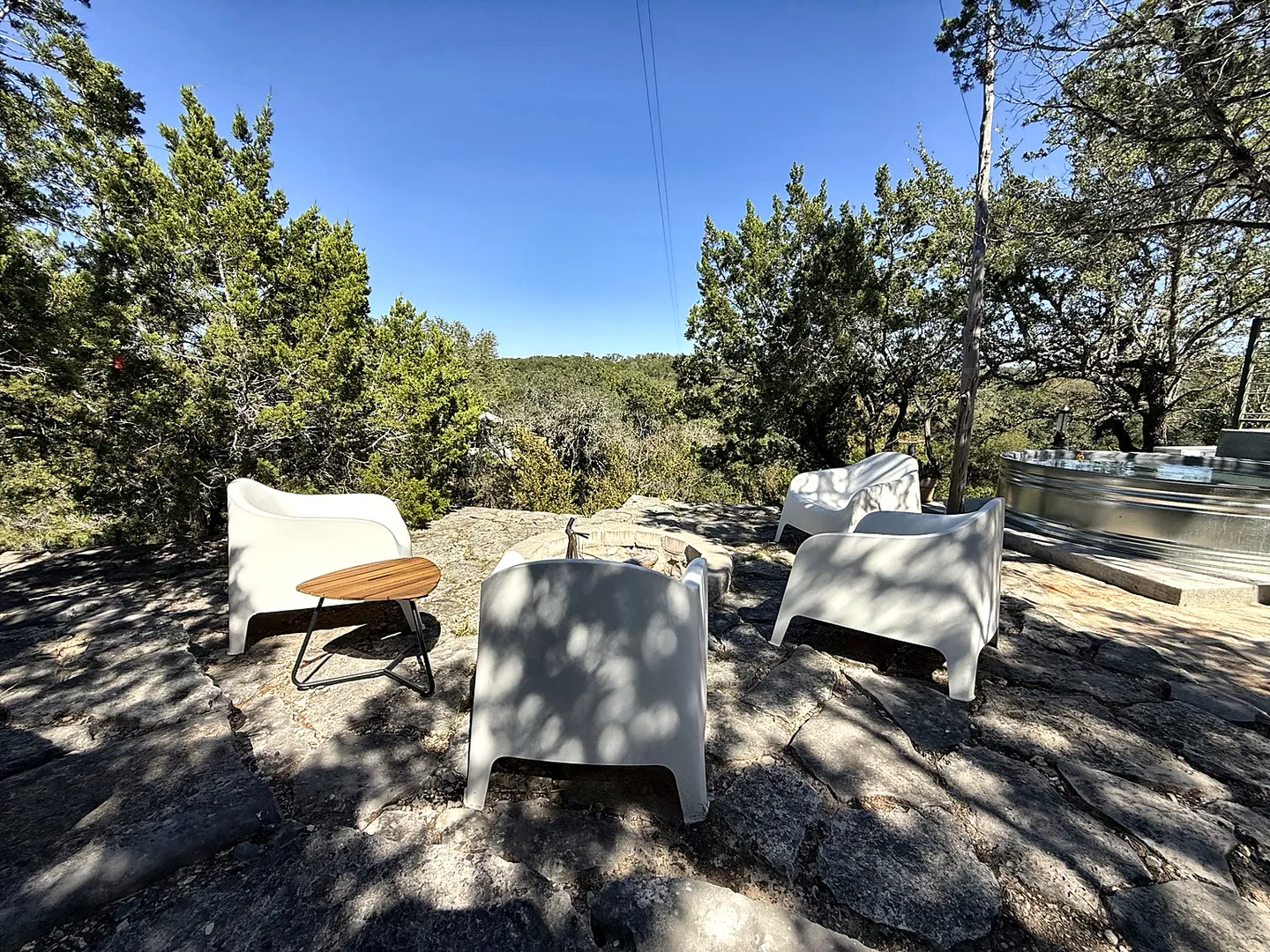 Outdoor stone patio with modern white armchairs surrounding a stone fire pit, overlooking green trees under a clear blue sky.