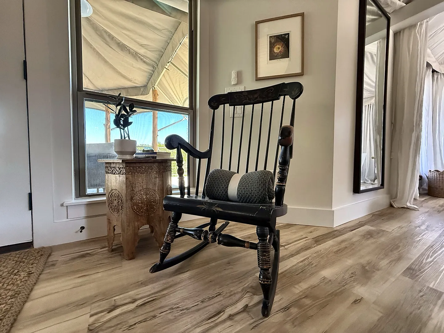 A black wooden rocking chair with gold accents and a rolled blanket set next to a window and carved side table.