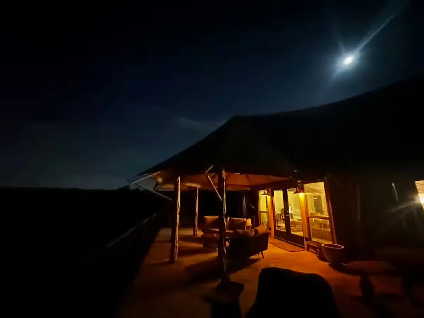 Covered outdoor deck with patio furniture illuminated by warm lights at night beneath a moonlit sky.