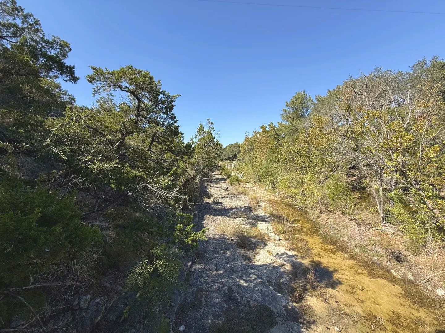 A dry, rocky creek bed with patches of shallow standing water flanked by trees and greenery under a clear blue sky.