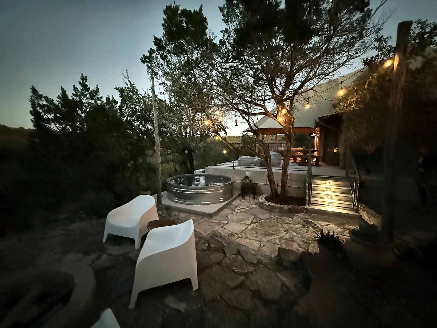 Dusk view of an outdoor stone patio with modern white chairs, a stock tank pool, illuminated deck steps, and string lights.