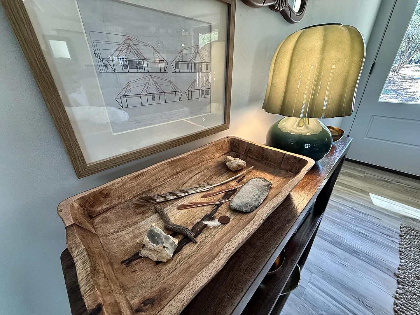 Close-up of a wooden console table featuring a carved wooden tray filled with natural elements, an illuminated lamp, and framed artwork.