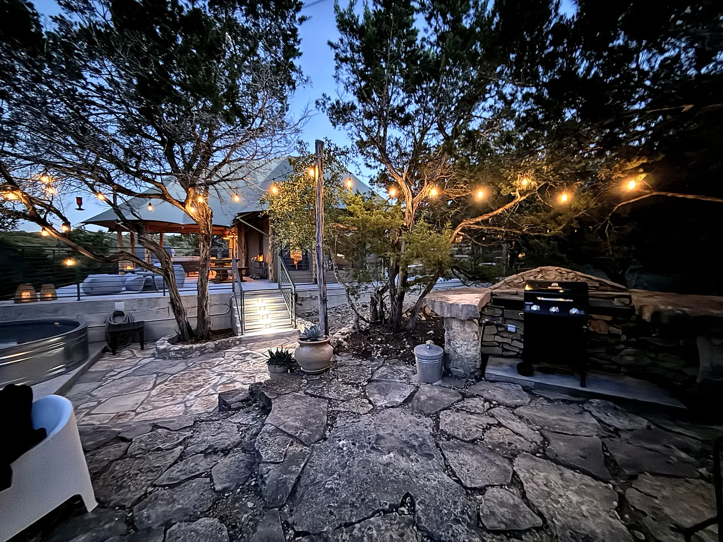 Outdoor stone patio at twilight with string lights, trees, a barbecue grill, and steps leading up to an illuminated safari-style tent structure.
