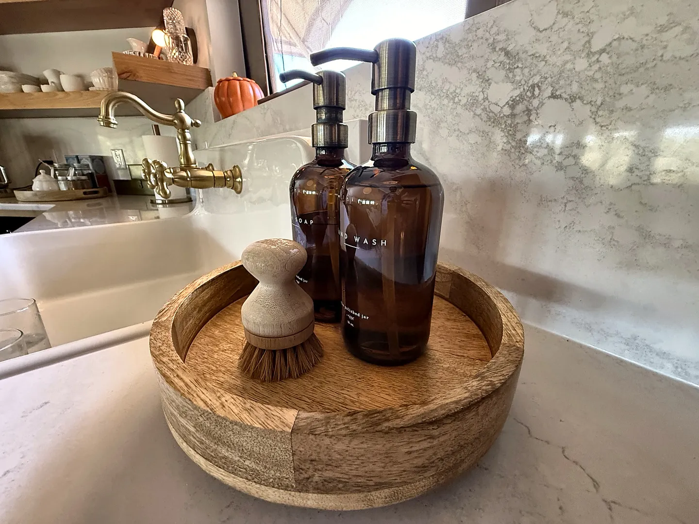Close-up of a round wooden tray on a countertop next to a sink, holding two amber glass pump bottles and a wooden scrub brush.