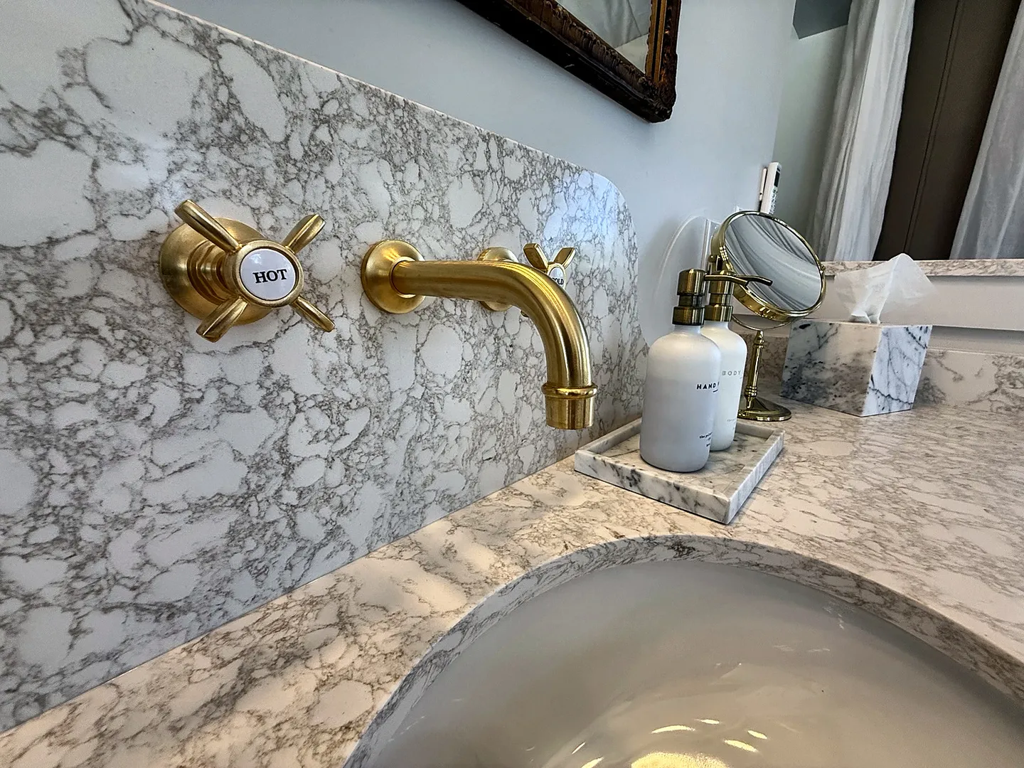 Close-up of a bathroom vanity featuring a wall-mounted brass faucet and handles against a marble-patterned backsplash and countertop.