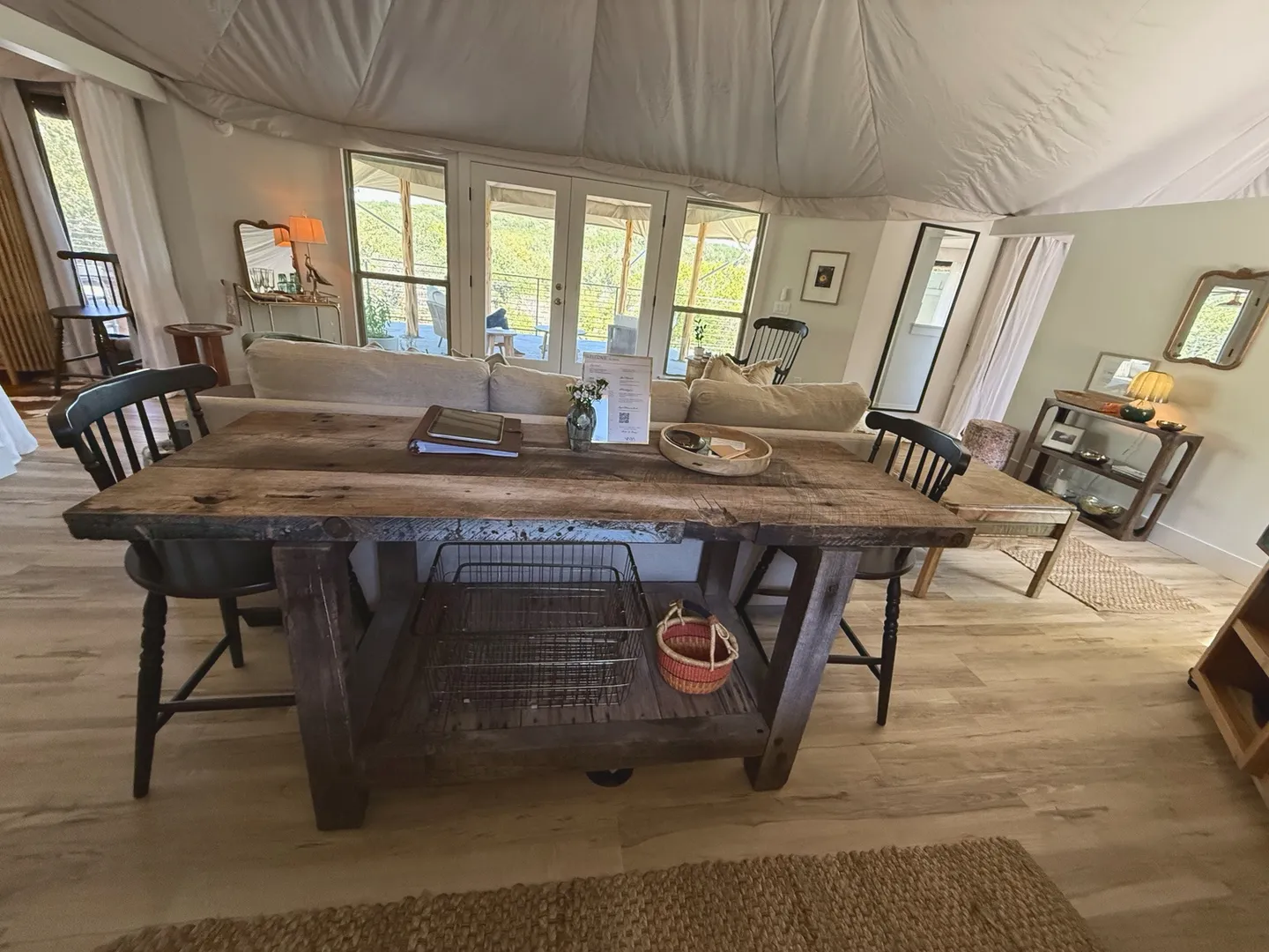 A rustic wooden dining table with black chairs situated behind a living room sofa under a tent-like ceiling.