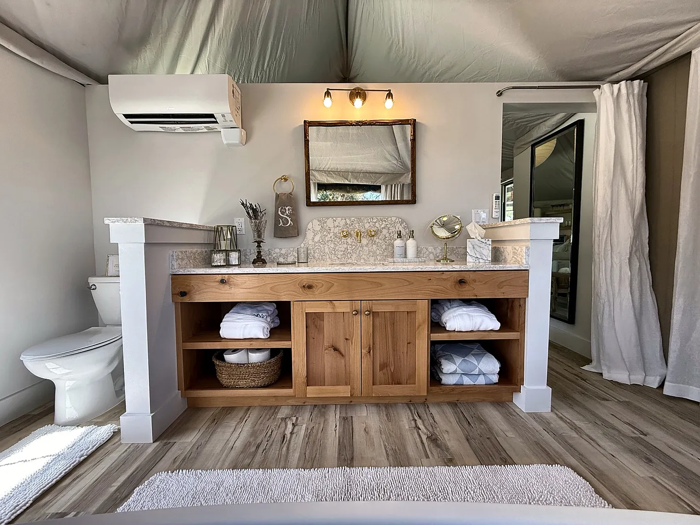 Bathroom vanity with a wooden base, integrated sink, and open shelving beneath a framed mirror inside a tent structure.