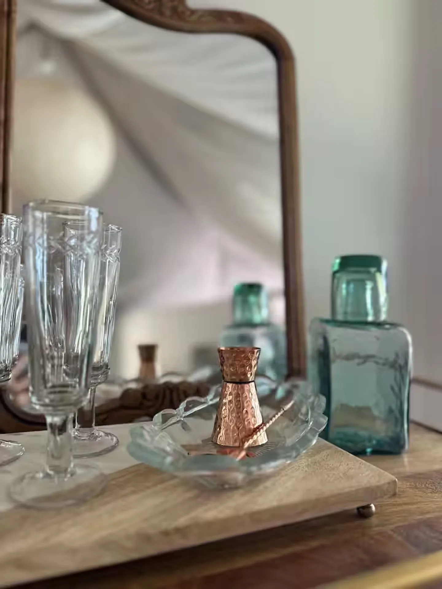 Close-up of glassware, a copper jigger in a glass dish, and a teal glass bottle in front of a mirror on a wooden tabletop.