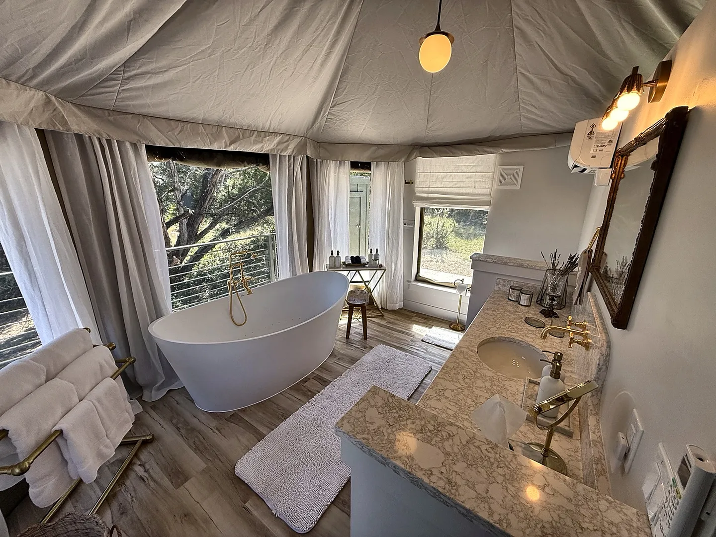 Bathroom inside a canvas tent structure featuring a freestanding oval soaking tub, large windows, and a vanity with marble-patterned countertops.