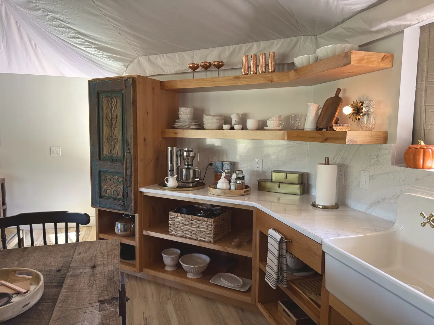 Kitchenette corner with wooden open shelving, light marble-style countertop, farmhouse sink, and tent-style fabric ceiling.