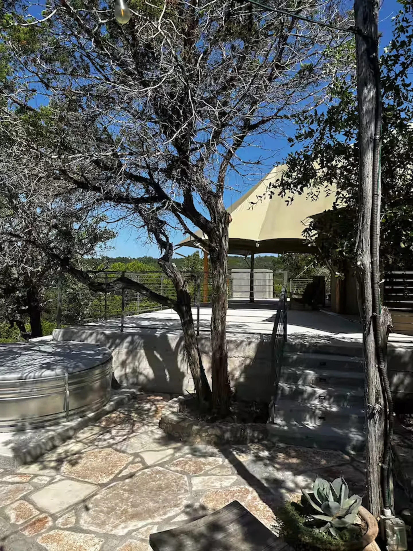 Outdoor stone patio with a metal stock tank pool, concrete steps leading to an elevated deck, and a canvas tent structure under trees and a blue sky.