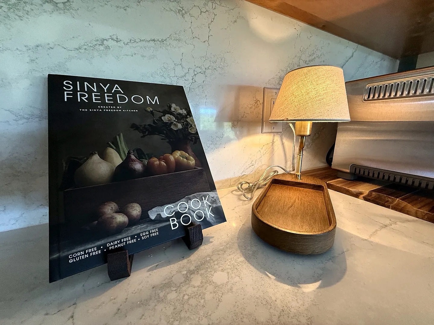 Close-up of a kitchen countertop featuring a displayed cookbook on a small stand next to a compact wooden base lamp.