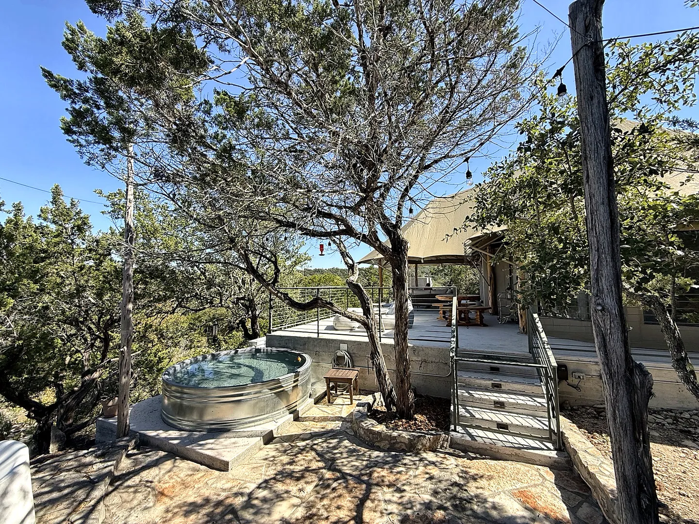 Outdoor area featuring a circular metal soaking tub next to a stone patio, mature tree, and elevated deck.