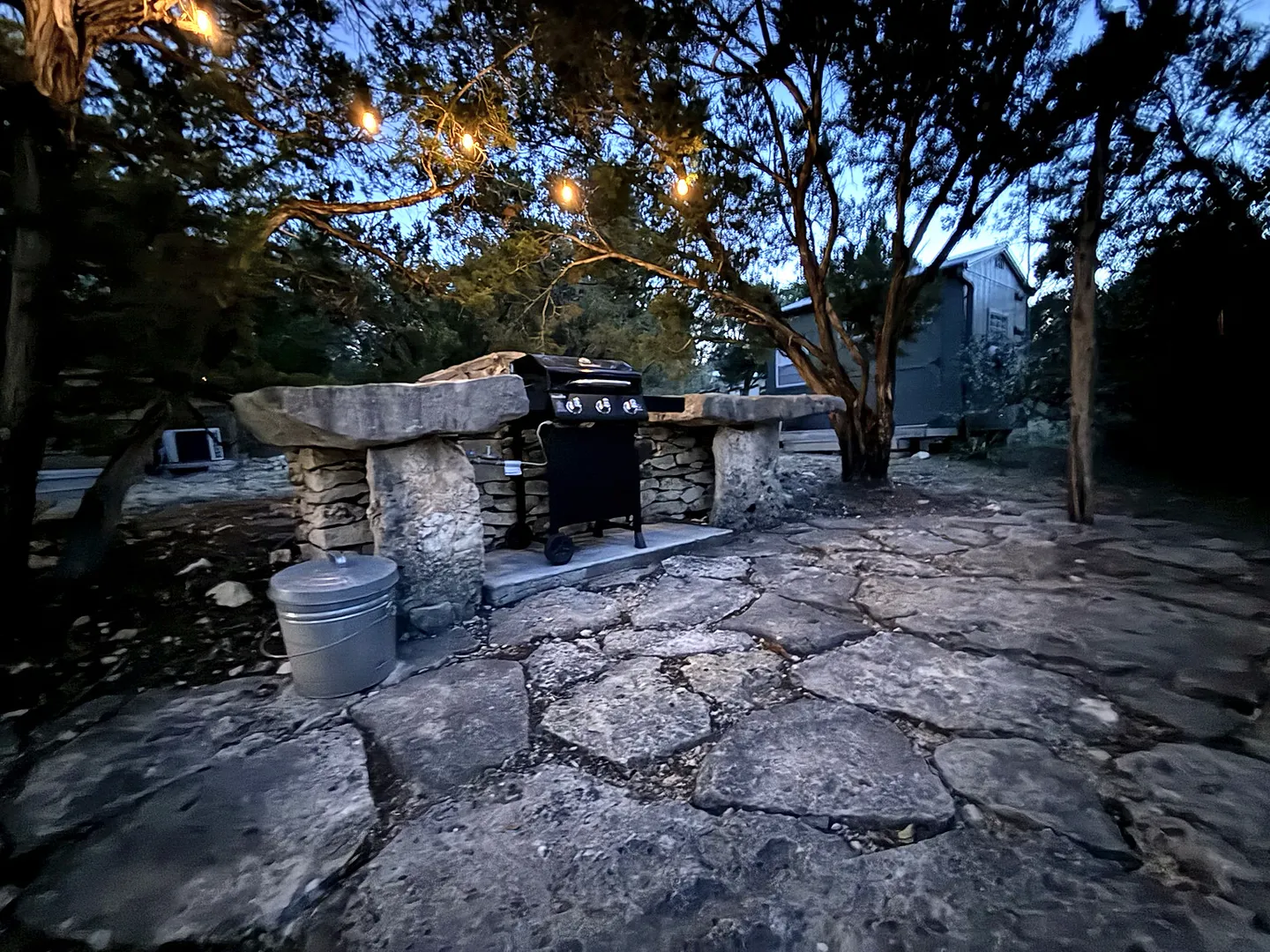 Outdoor barbecue area with a stone-slab counter, gas grill, stone patio, and string lights hung among trees at dusk.