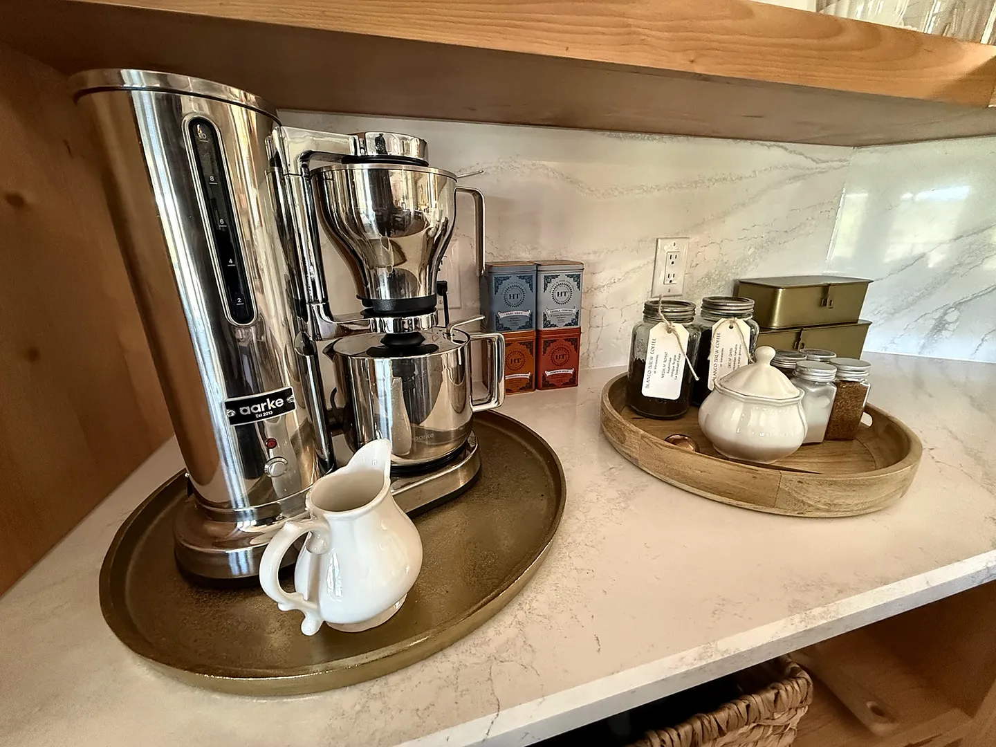 Close-up of a stainless steel coffee maker on a countertop tray with a cream pitcher, alongside jars and tea tins.