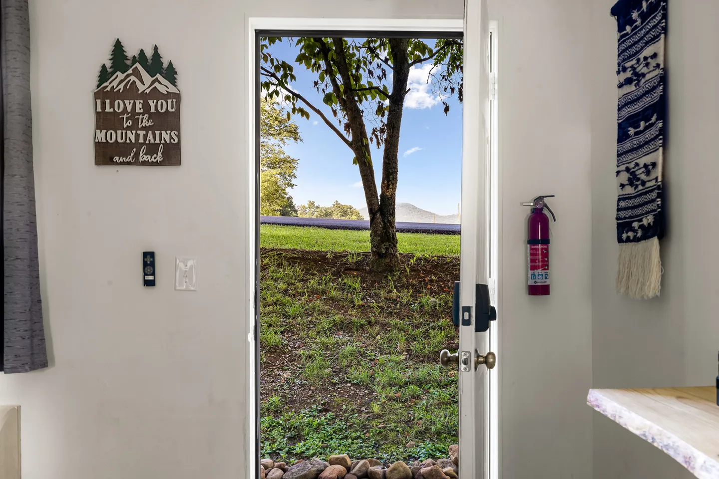 Open doorway looking out toward a grassy yard with trees, a paved path, and mountains under a blue sky.