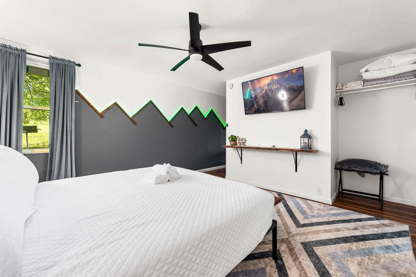 Bedroom featuring a bed with white bedding, a ceiling fan, a wall-mounted TV, and an accent wall with mountain-shaped molding and green backlighting.