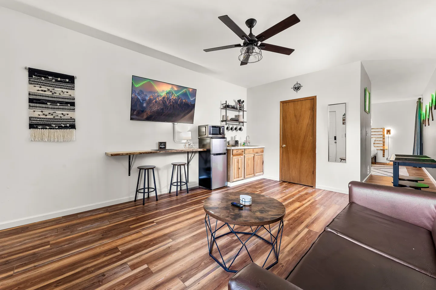 Living area with wood-look flooring, a wall-mounted TV, a bar counter with stools, a kitchenette nook, and a sofa.