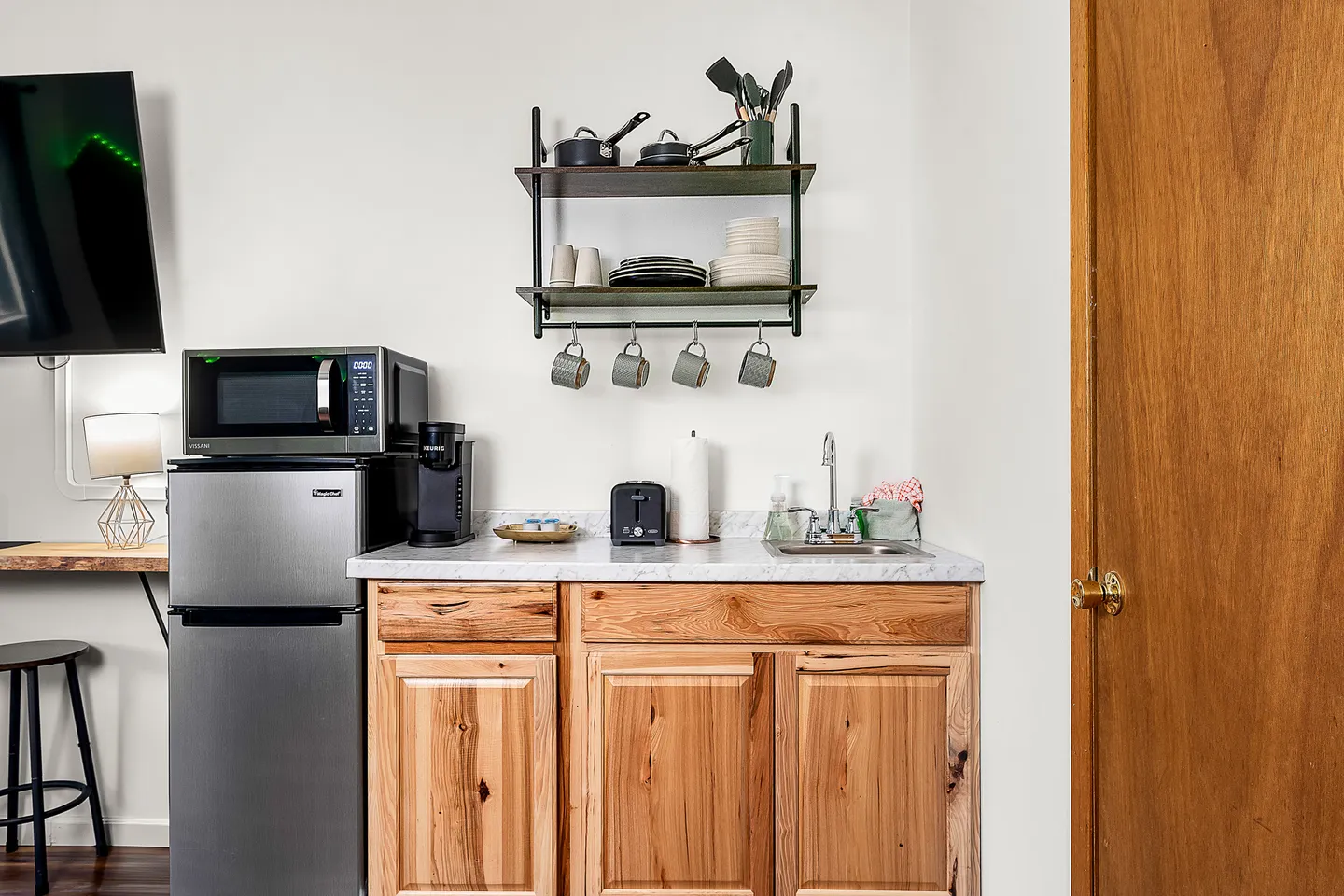 A kitchenette setup featuring natural wood base cabinets, a light marble-look countertop, mini fridge, microwave, sink, and hanging wall shelves with cookware.