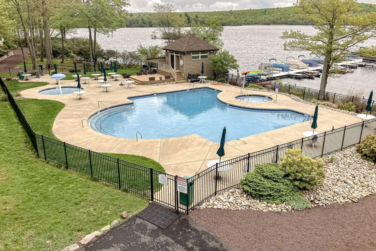 Elevated view of a fenced outdoor swimming pool complex overlooking a lake, featuring a main pool, hot tub, kiddie pool, and boat docks.
