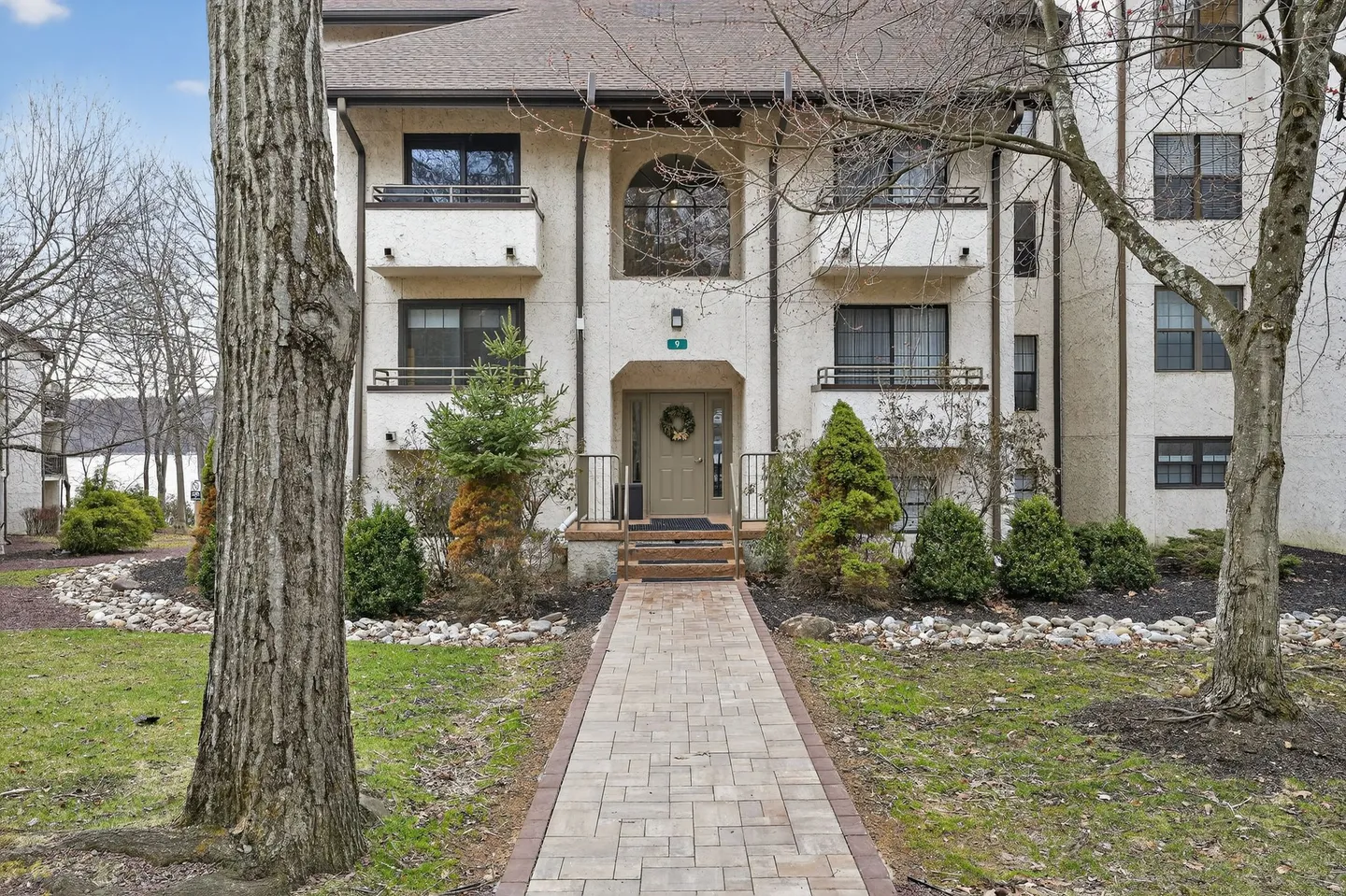 Paved walkway leading to the ground-floor entrance of a multi-story stucco building with balconies and surrounding trees.