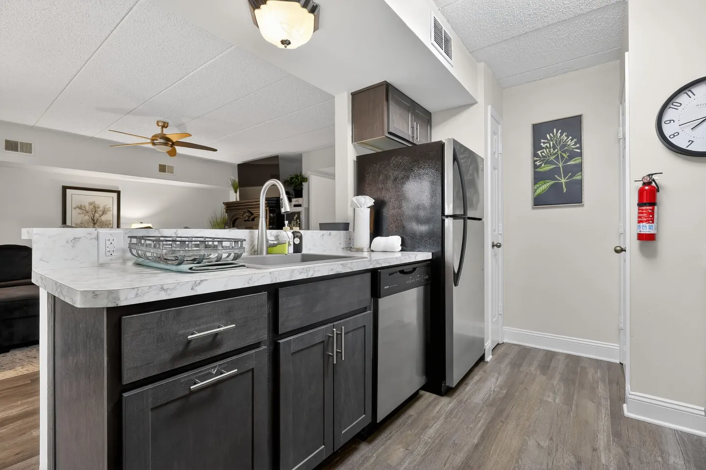 A kitchen with dark cabinetry, stainless steel dishwasher and refrigerator, and light countertops overlooking an adjacent living area.