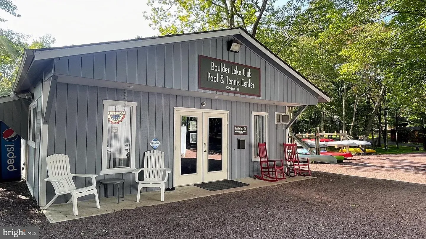 Exterior of the Boulder Lake Club Pool and Tennis Center check-in building with white and red armchairs out front and kayaks stored in the background.