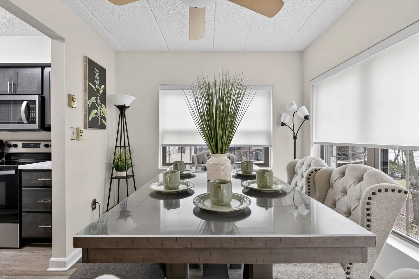 Dining room table set with plates and mugs, flanked by tufted upholstered chairs and windows with white shades.