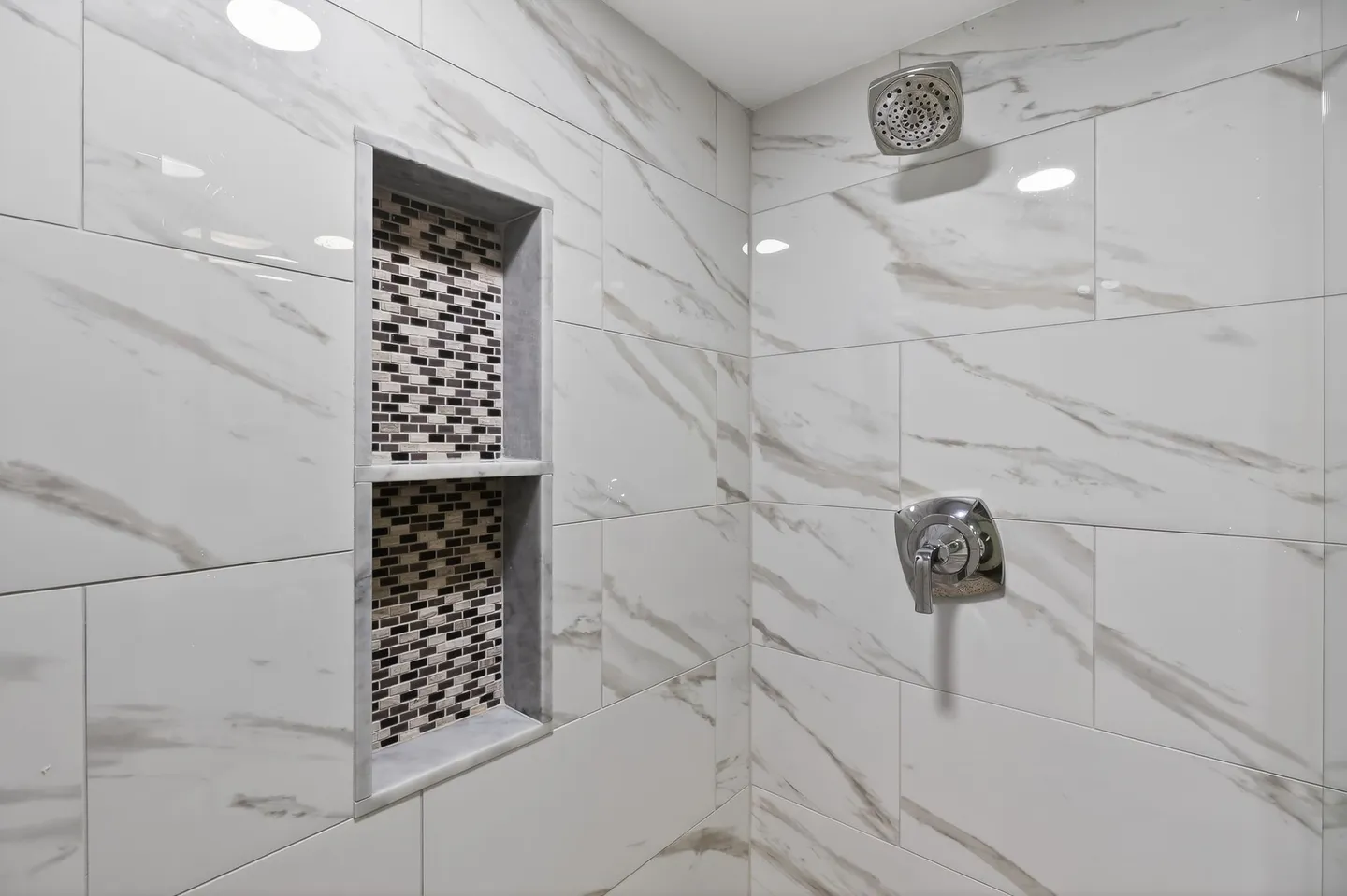 Close-up of a tiled shower stall with white marble-pattern tiles, chrome fixtures, and a recessed two-tier niche with mosaic tile backing.
