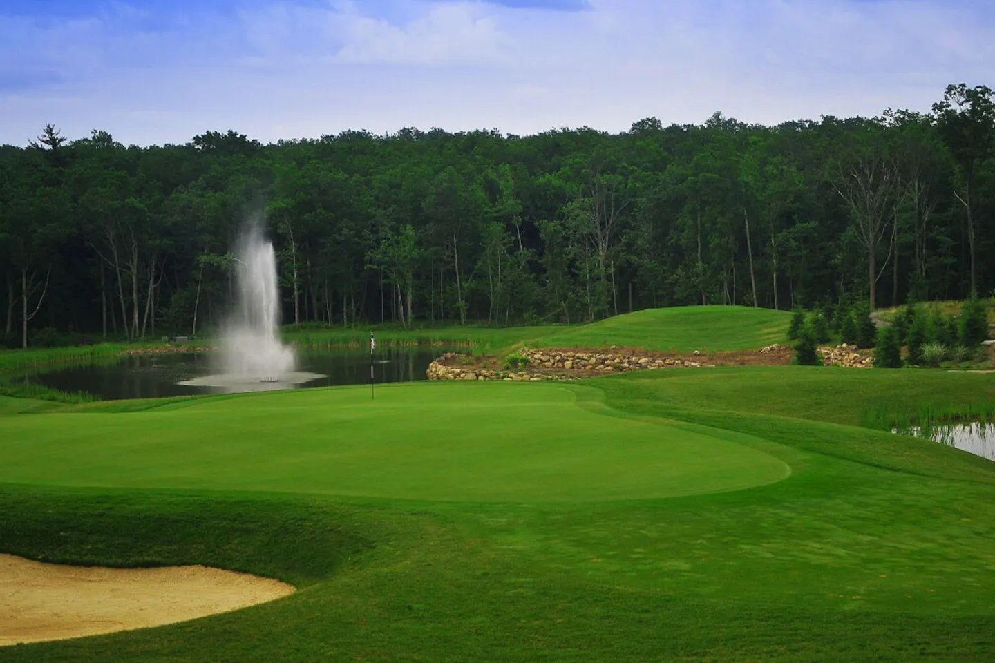 A golf course putting green bordering a pond with a spraying water fountain and dense forest behind.