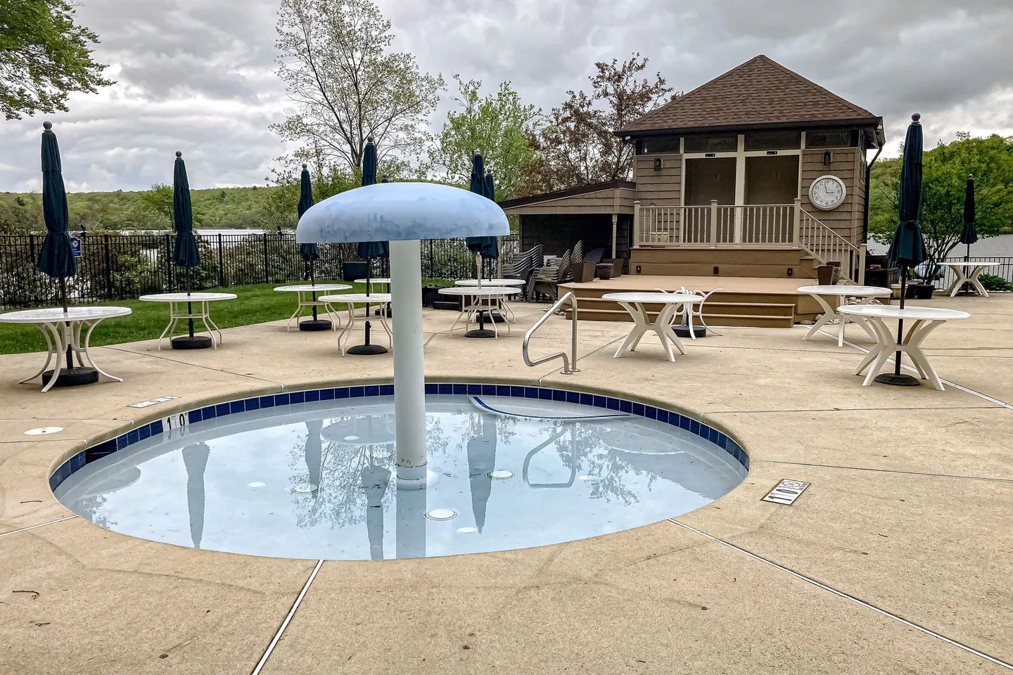 A shallow circular wading pool with a blue mushroom fountain on a concrete deck surrounded by outdoor tables and a pool house.