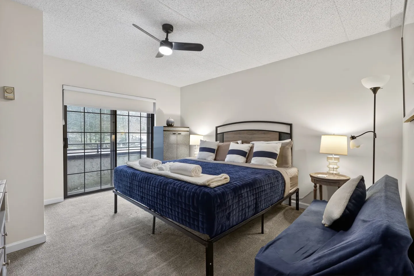 Bedroom with a bed made with blue bedding, a blue futon couch, and sliding glass doors leading outside.