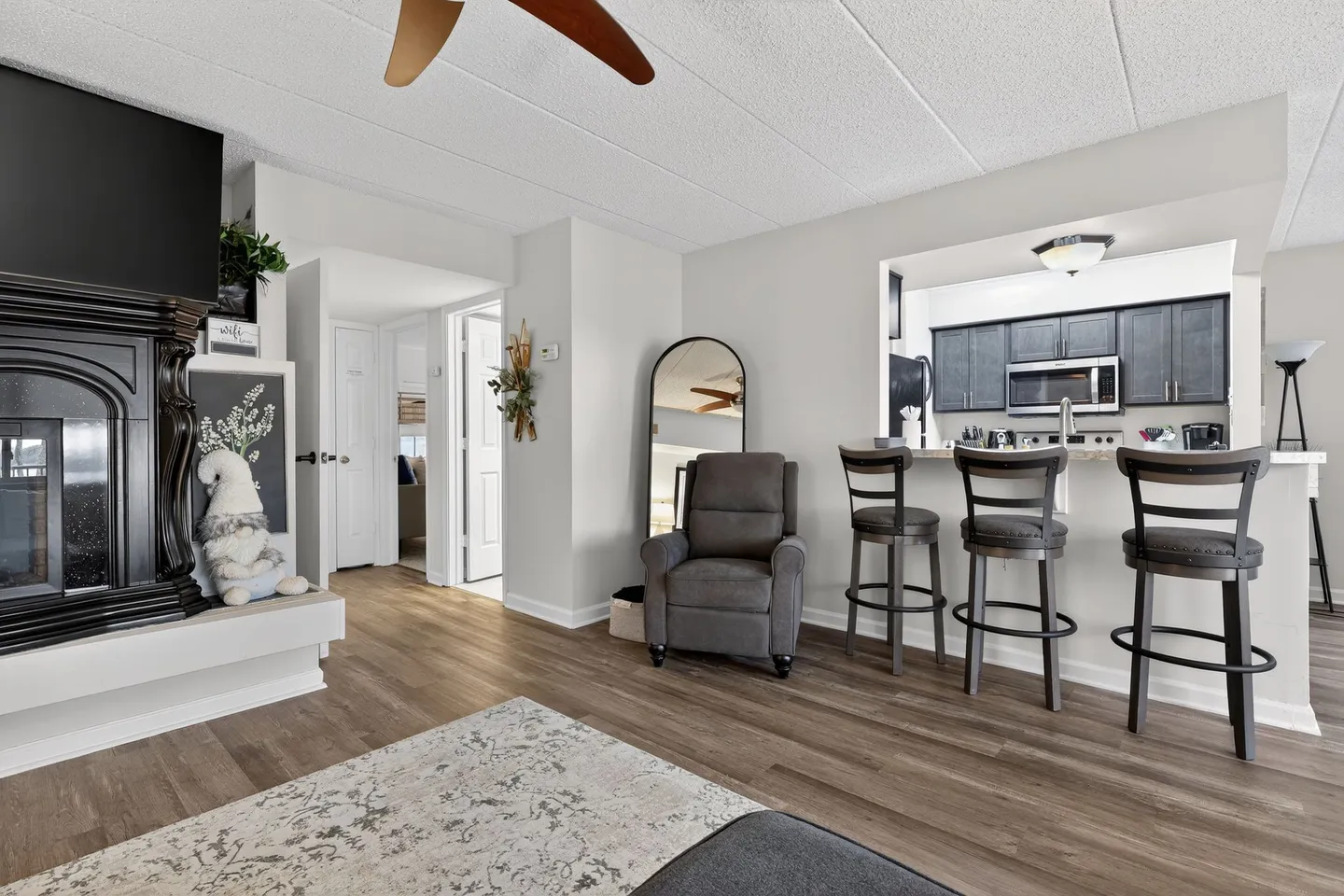 Living area with wood-look flooring, a fireplace on the left, an armchair, and a kitchen breakfast bar with three stools.