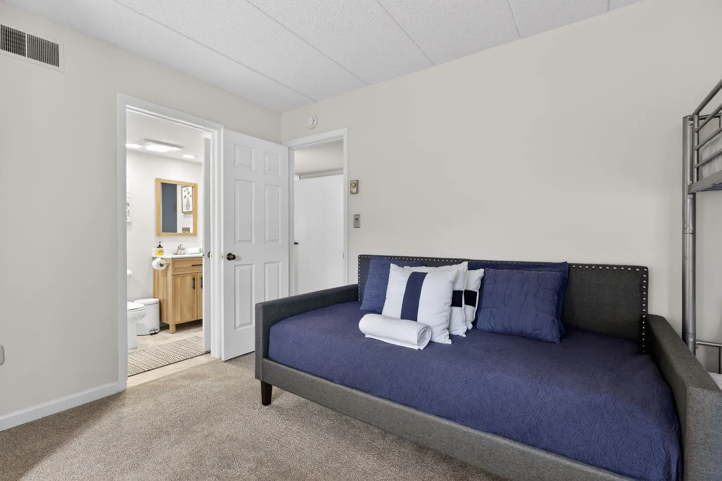 Bedroom with carpeted flooring, a daybed with blue bedding, and an open doorway leading to an ensuite bathroom.