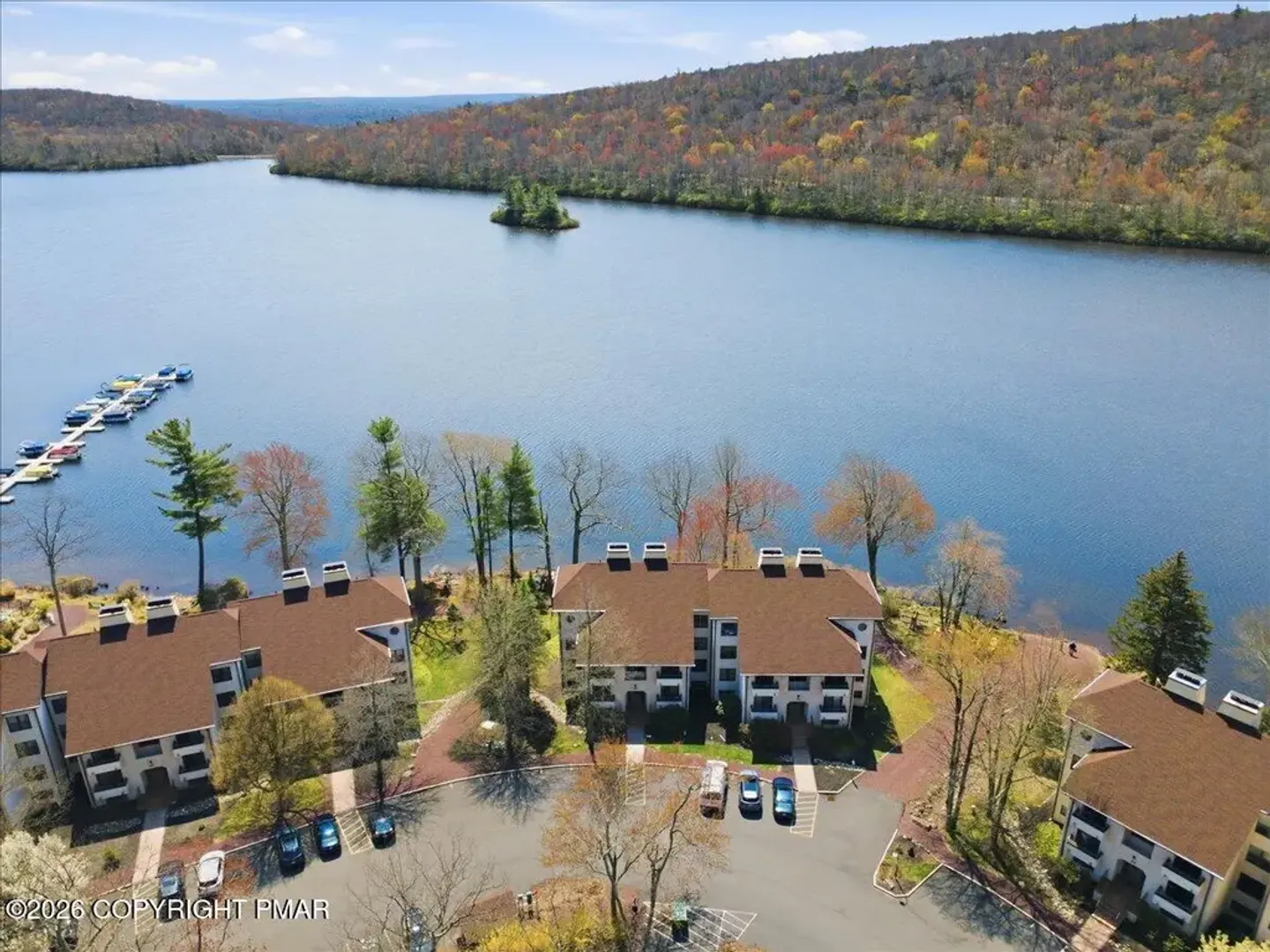Aerial view of a lakefront residential community with multi-story buildings, parking areas, boat docks, and forested hills across the water.