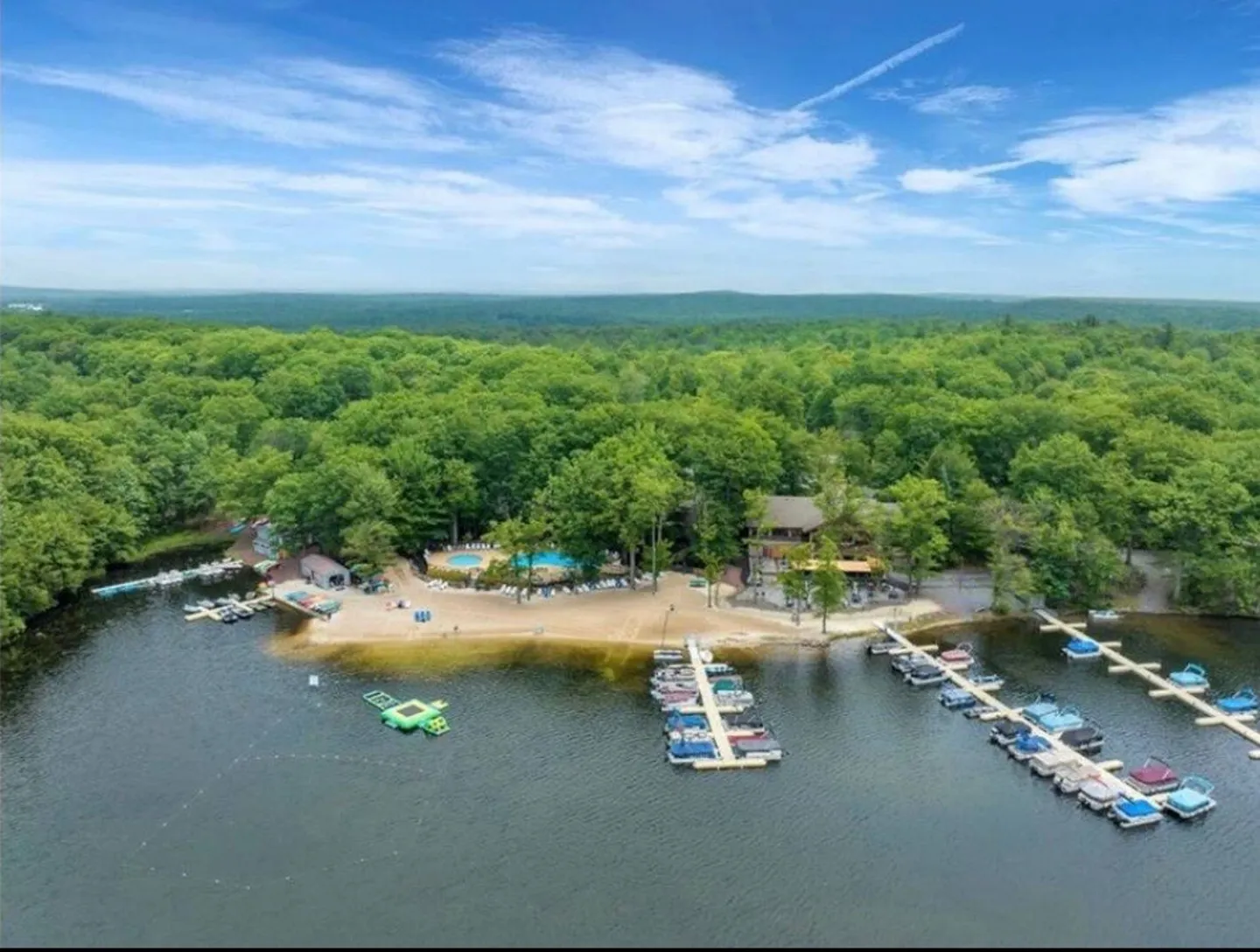 Aerial view of a lakefront resort with a sandy beach, boat docks, an outdoor swimming pool, and dense forest surroundings under a blue sky.