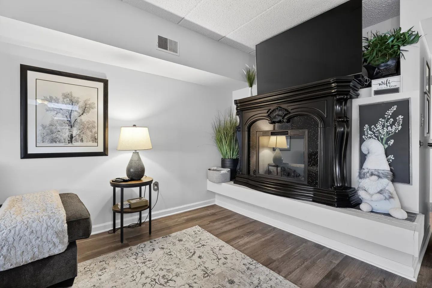 Living room corner featuring an ornate black fireplace mantel on an elevated white hearth beneath a wall-mounted TV.