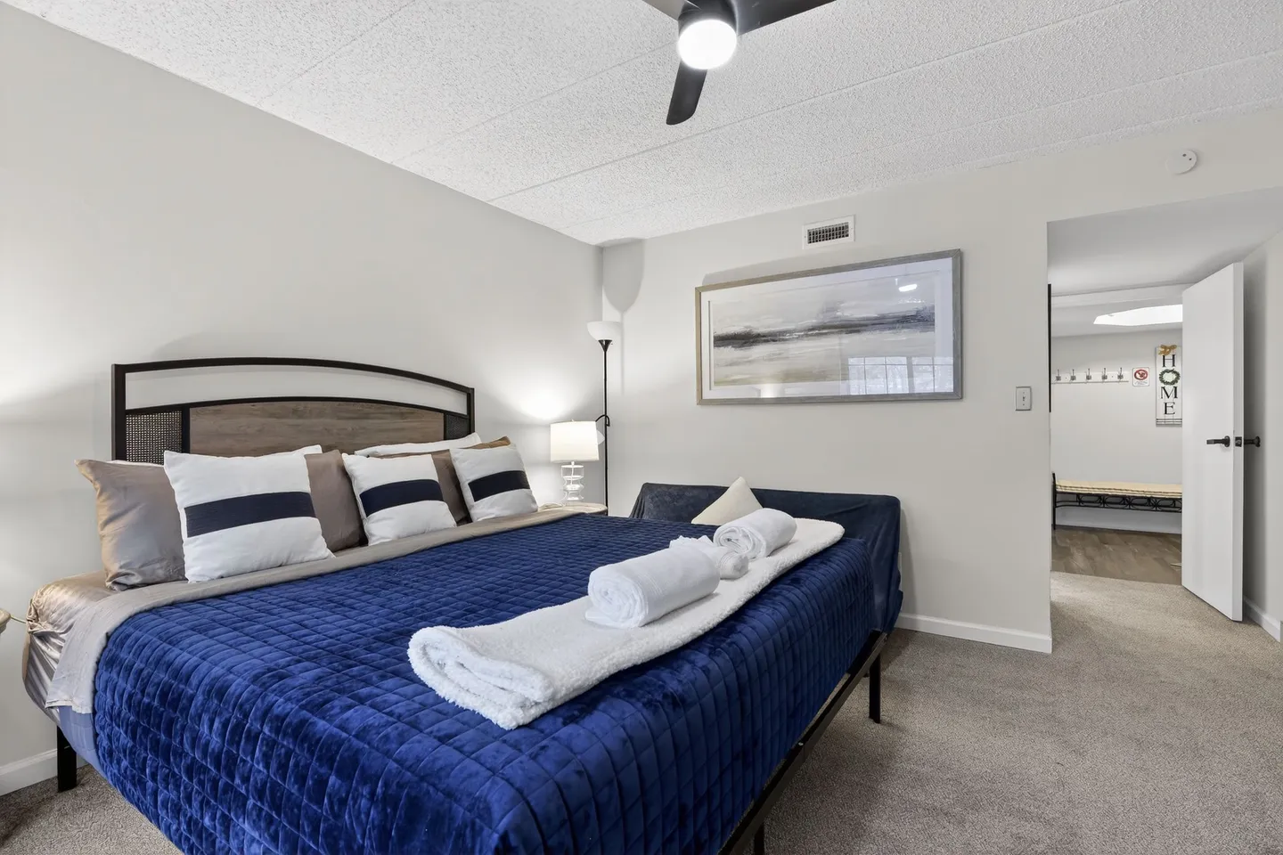 Bedroom featuring a made bed with a blue quilted coverlet, white folded towels, and an open doorway leading to a hallway.