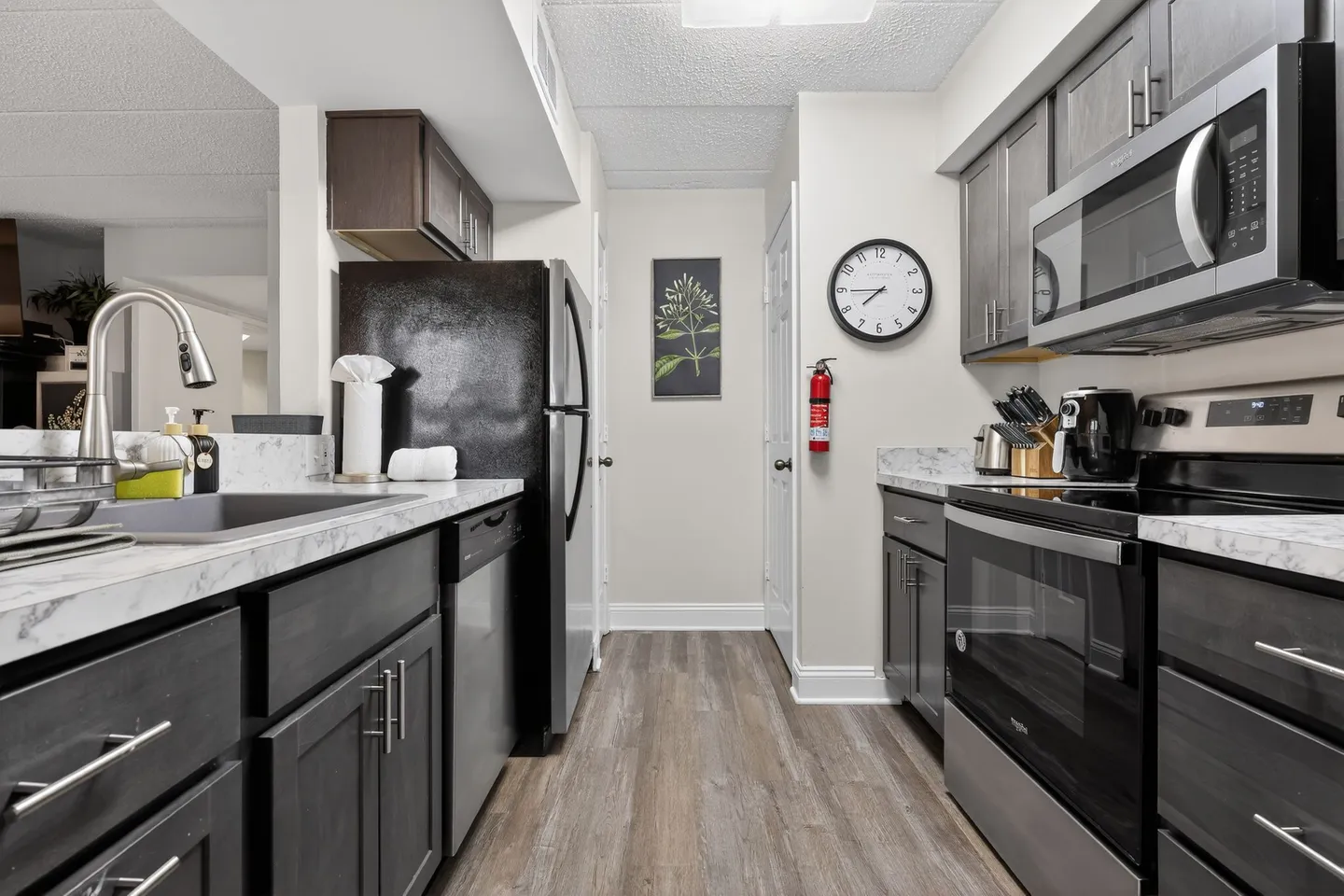 Galley kitchen with dark wood-grain cabinets, light marbled countertops, black and stainless steel appliances, and wood-look flooring.