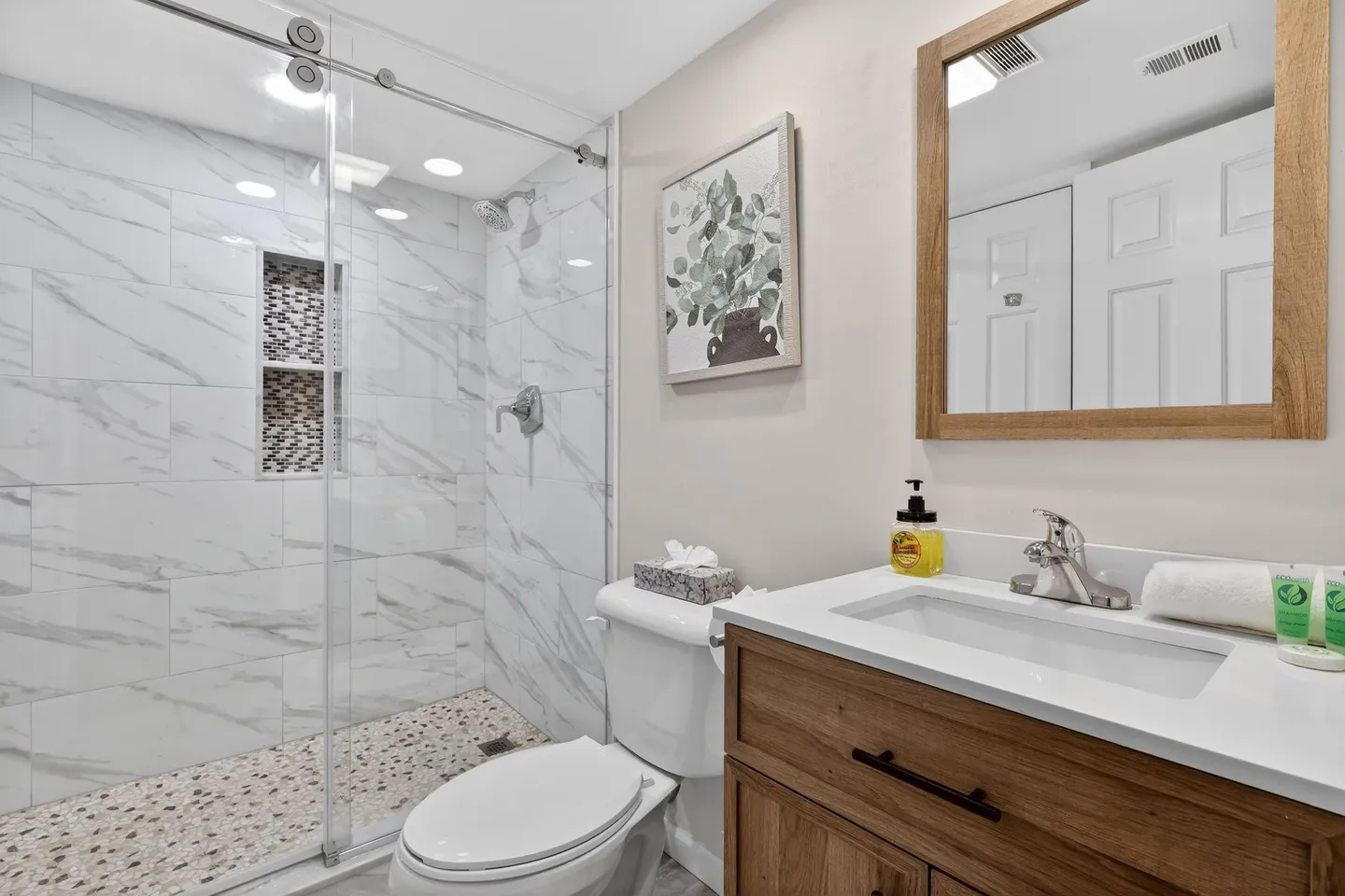 Bathroom with a glass-enclosed walk-in shower featuring marble-patterned tile, pebble flooring, a toilet, and a wood-toned vanity with a framed mirror.