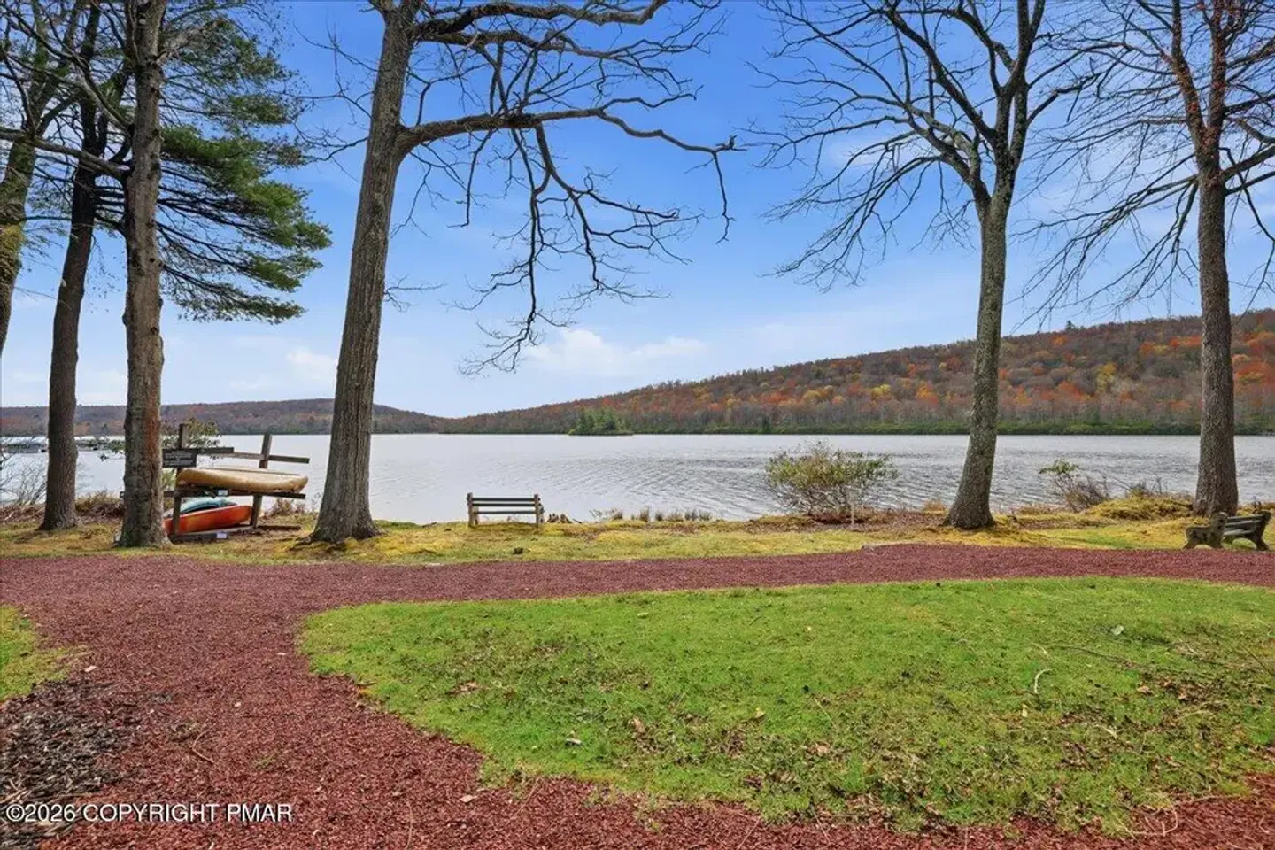 A grassy lakefront yard with a red gravel path, mature trees, a wooden bench, and canoes stored near the water edge looking across to forested hills.