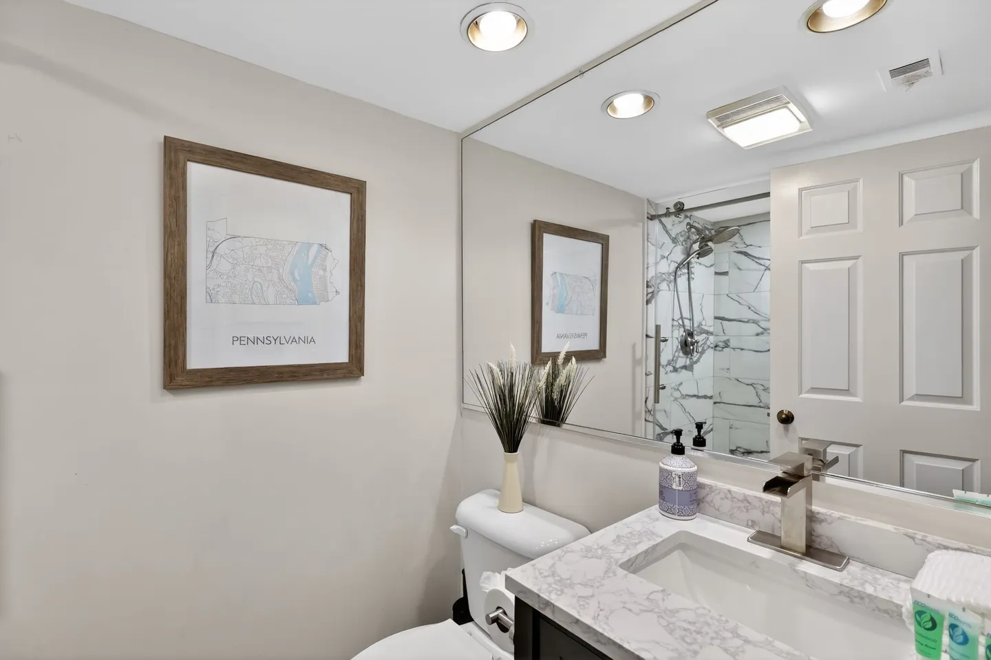 Bathroom with marble-patterned countertop, toilet, large wall mirror, framed Pennsylvania map, and reflection of a walk-in shower with marble-look tile.
