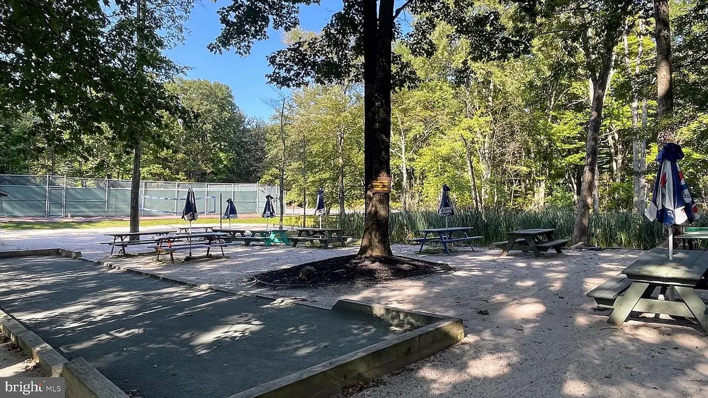 Outdoor recreation area with a bocce court, picnic tables with umbrellas, and tennis courts in the background surrounded by trees.