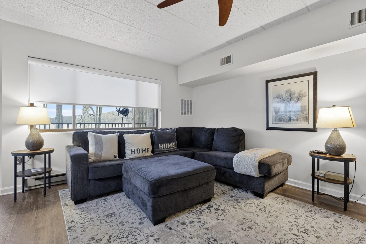 Living area featuring a dark gray sectional sofa, matching ottoman, patterned area rug, and end tables with lamps beside a window.