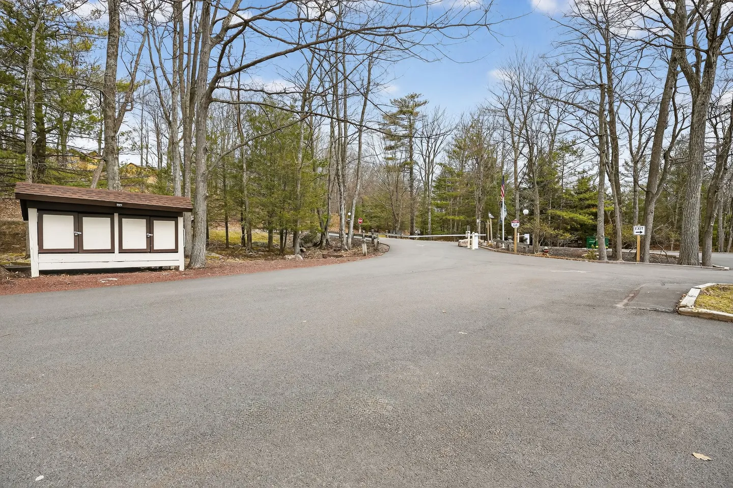 Paved entrance road surrounded by trees with a gated checkpoint barrier and a small mailbox or utility shed on the left.